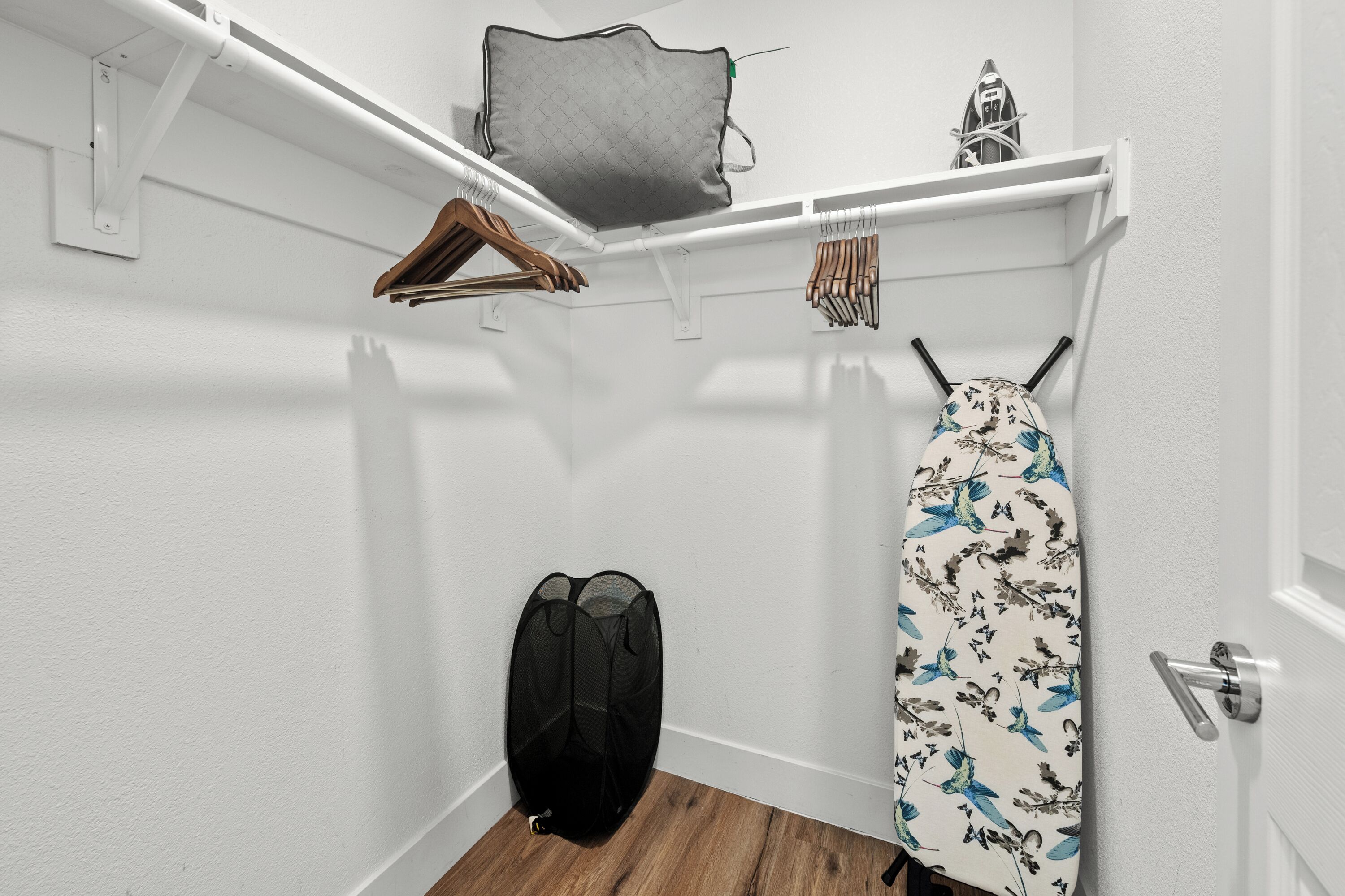 A corner of a room with a wooden floor, a hanging garment bag, a shelf with a grey bag and a decorative item, and a door with a silver handle.