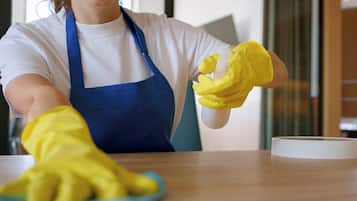 Una persona con delantal azul y guantes amarillos sostiene un pulverizador, probablemente limpiando una mesa.