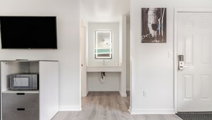 A modern hotel room with a flat-screen TV, a microwave, a built-in sink with a mirror, and a door with a wine glass artwork. - Blue Beach Motel (North Kingstown)