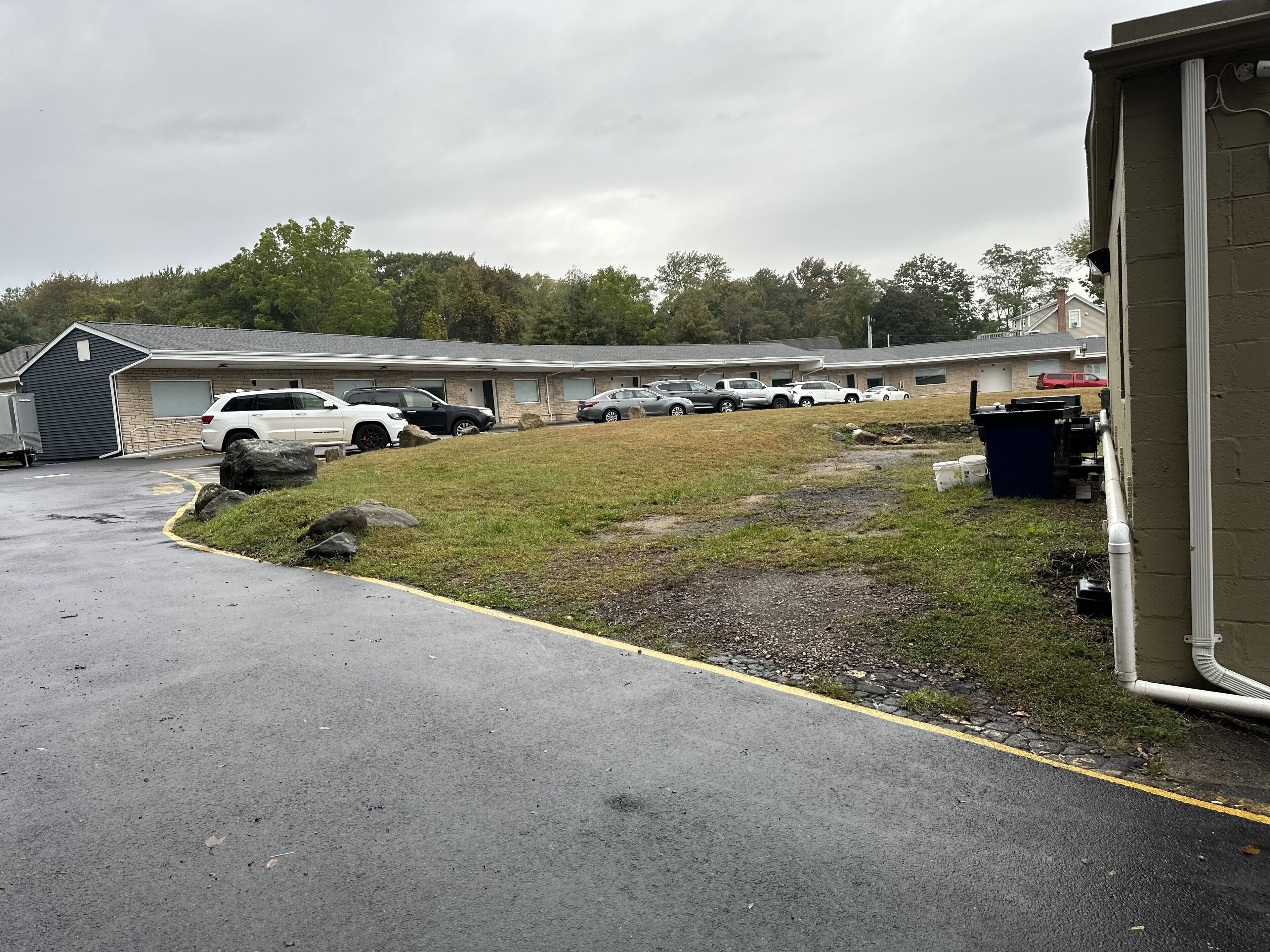 A parking lot with several cars, a building with a garage, and a grassy area with rocks.
