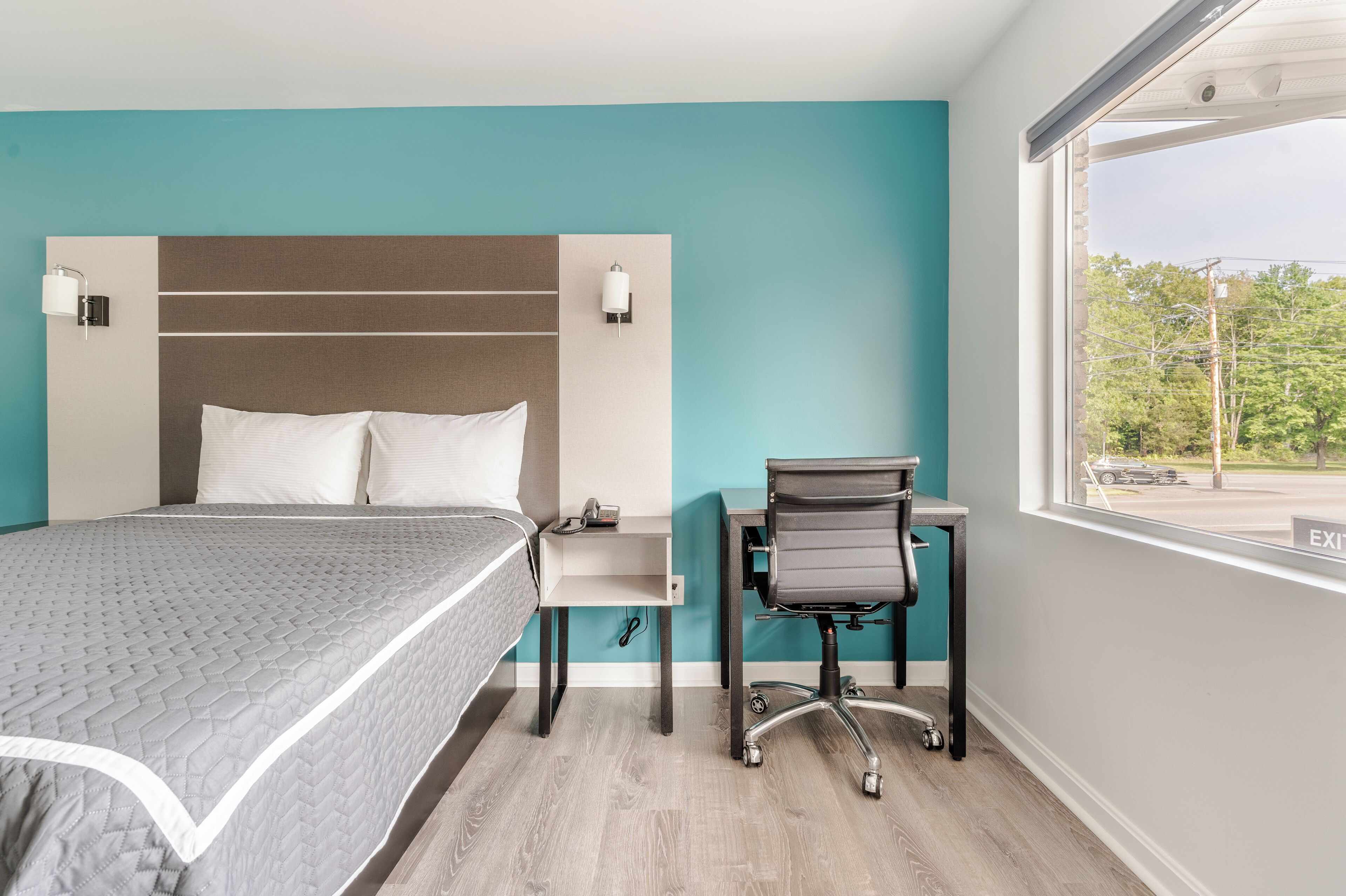 A hotel room with a bed, a desk, and a window with an exit sign.
