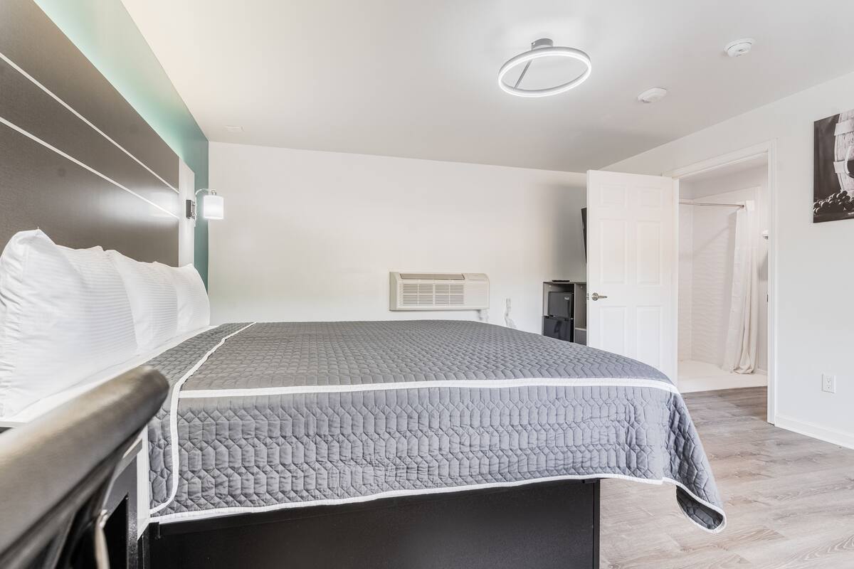 A bedroom with a bed, a bedside table, a wall-mounted air conditioner, and a door leading to another room.