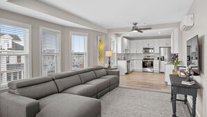 A modern living room with a sectional sofa, a dining area, and a kitchen with white cabinets and stainless steel appliances.
