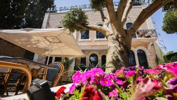An outdoor seating area with wicker chairs and a large umbrella, a tree, and a building with a sign reading "ALL SEASONS HOTEL".