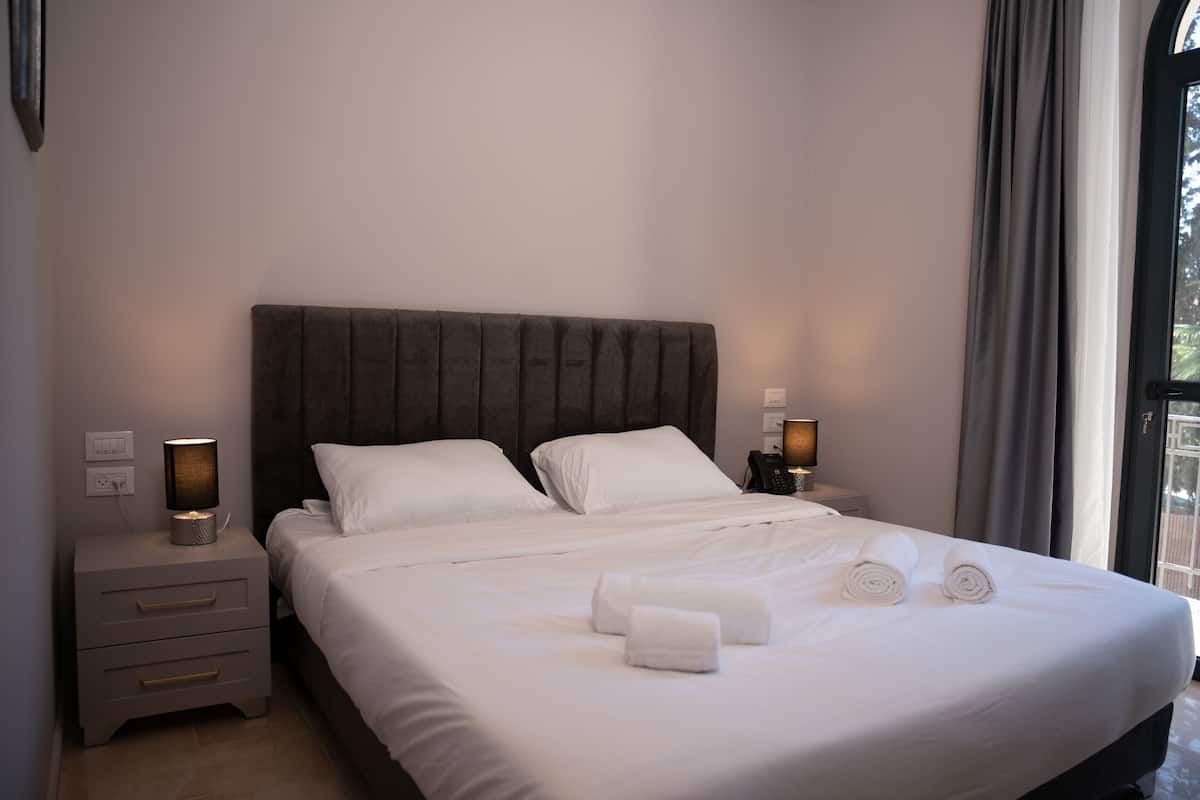 A neatly made bed with white linens and two rolled towels, a bedside lamp, and a nightstand with drawers.
