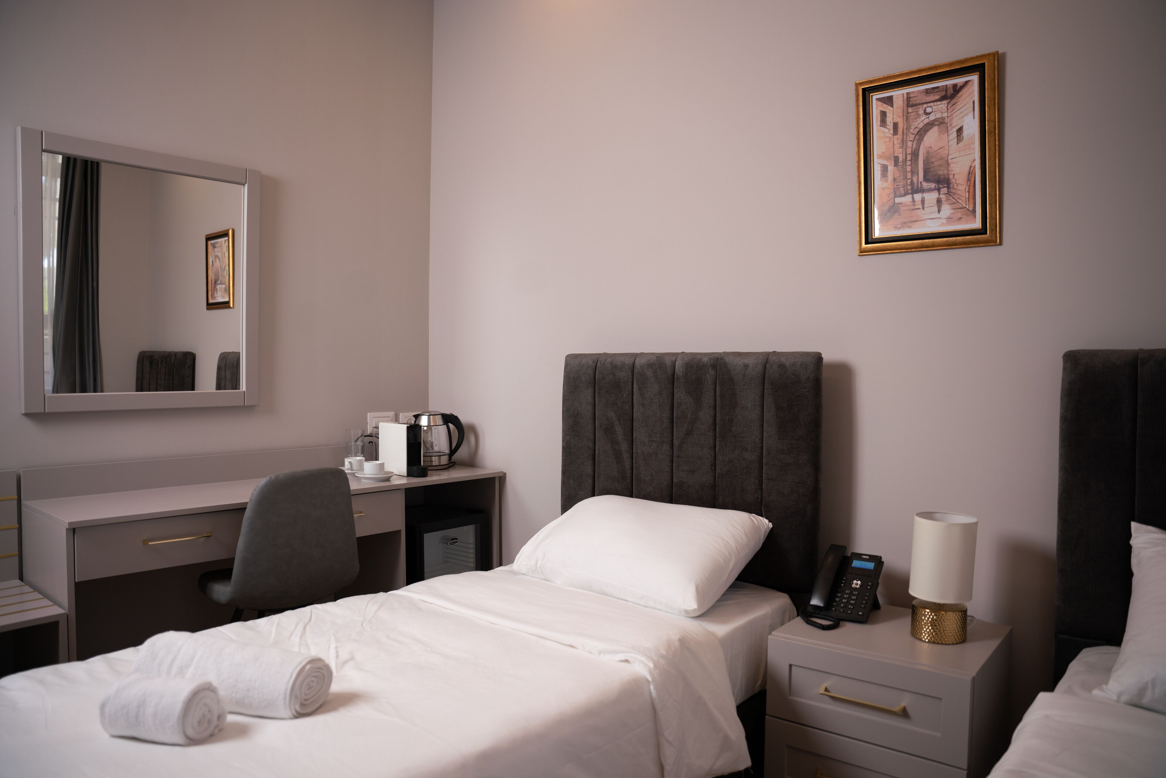 Quarto de hotel com cama, escrivaninha, cadeira, mesa de cabeceira com abajur, telefone e quadro na parede.