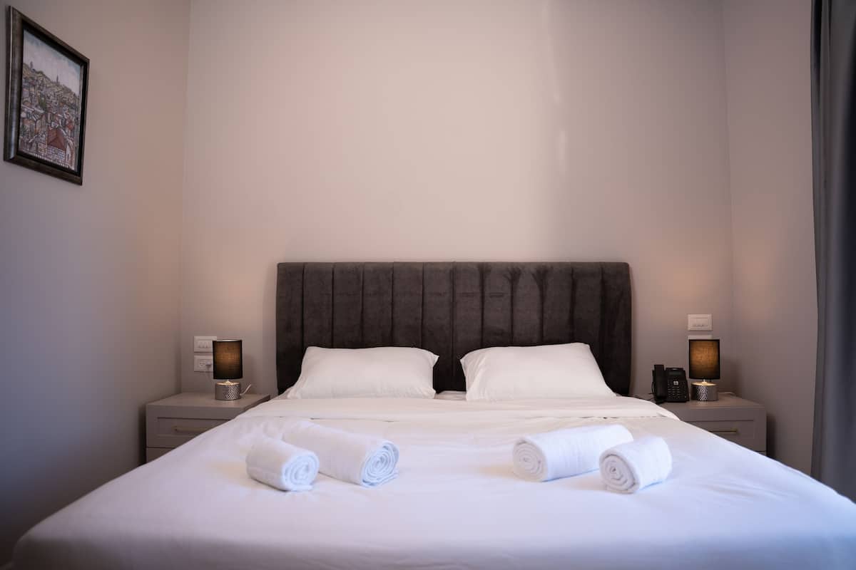A neatly made bed with white linens and two rolled towels, a dark headboard, bedside lamps, and a framed picture on the wall.