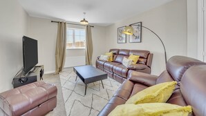 Un salon avec un canapé en cuir marron, un pouf noir, un téléviseur à écran plat et une lampe sur pied.