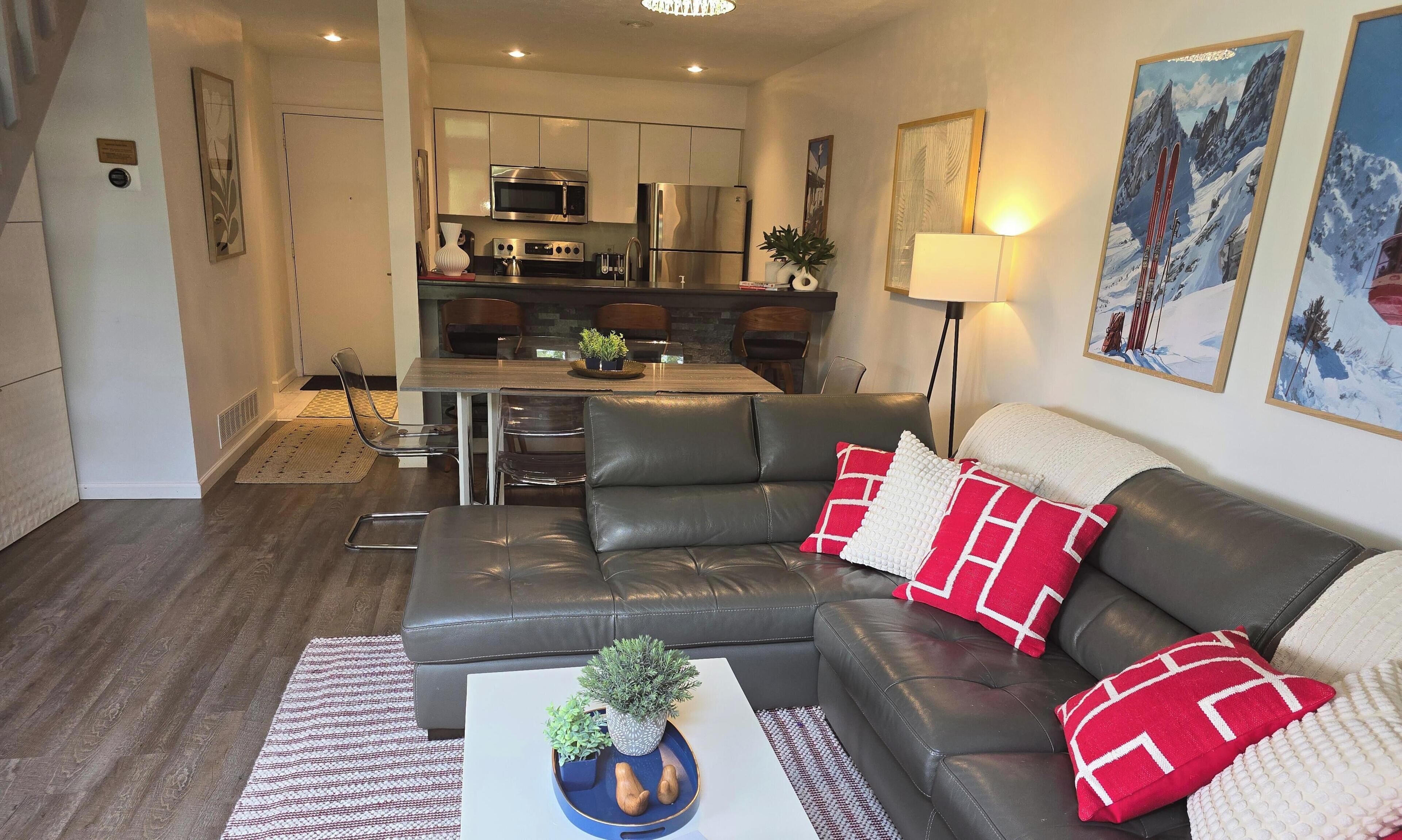 A modern living room with a leather sofa, a coffee table with a plant, and a dining area with a kitchen in the background.