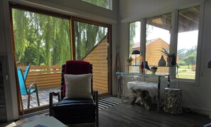 Ein Zimmer mit einer großen Schiebeglasdoor, die auf eine Außenterrasse führt, ein Sessel mit roter Decke, ein Glasregal mit Dekoration und ein weißer, flauschiger Hocker.