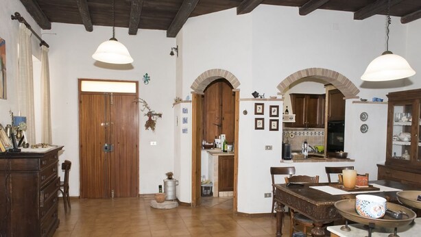 Ein geräumiges Zimmer mit Holzboden, einer großen Holztür, einem Esstisch mit Stühlen und einem durch einen Bogen sichtbaren Küchenbereich.