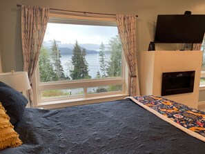A bedroom with a large window, a bed with a patterned quilt, a television mounted on the wall, and a fireplace.