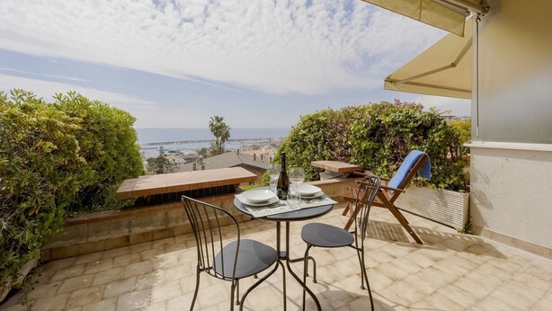 A terrace with a table set for two, chairs, a bottle, and glasses. - GuestHost - Amazing studio of 36 sqm, able to accommodate up to 3 people, located on the ground floor of a building in the quiet residential complex of Collina Fiorita. The property boasts a spacious terrace with fabulous views of Porto Sole and the avai (Sanremo)