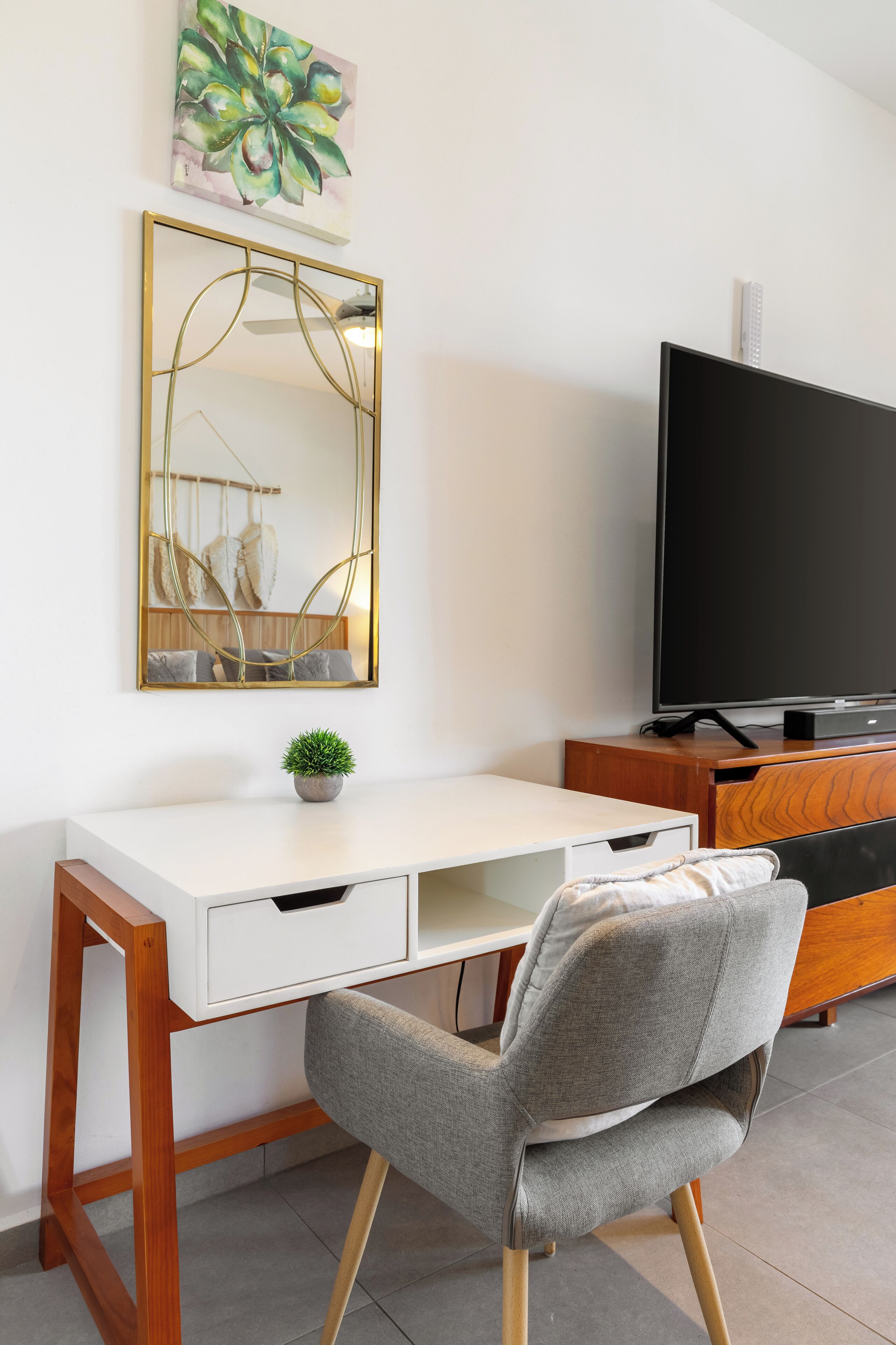 A modern room with a white desk, a grey upholstered chair, a large flat-screen TV, a wooden cabinet, and a wall-mounted mirror.