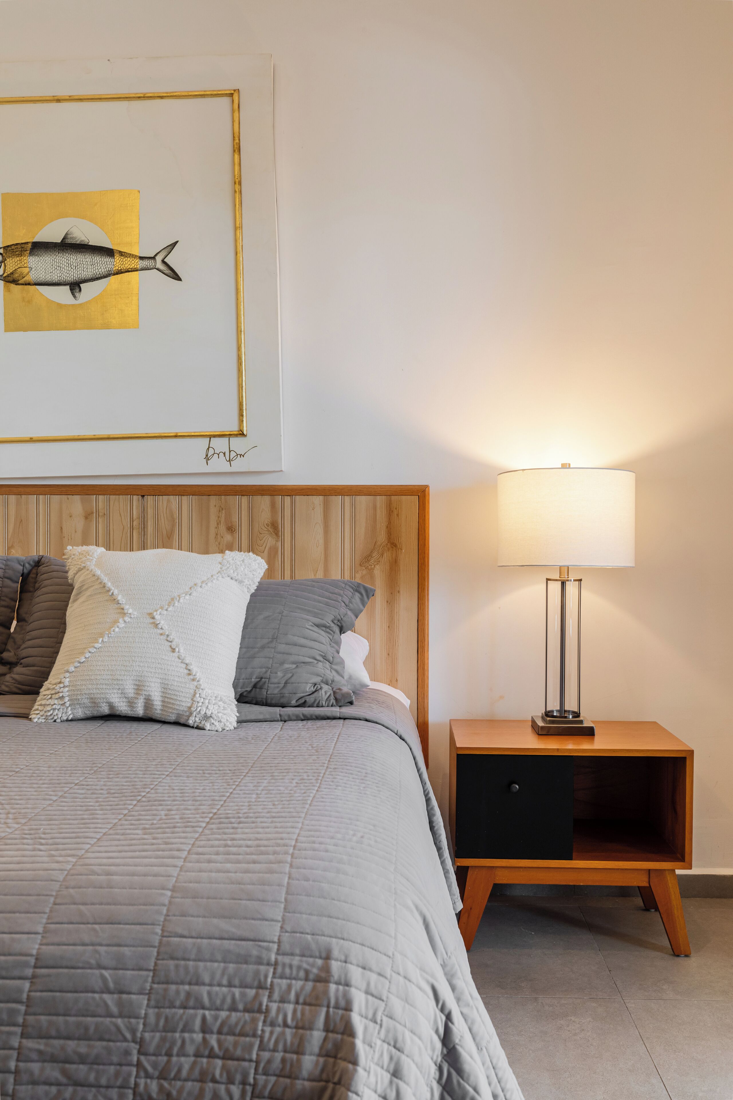 A neatly made bed with a grey quilt, two pillows, a wooden headboard, a bedside table with a lamp, and a framed artwork above the bed.