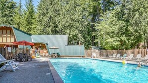 Une piscine dotée de chaises longues et d’un espace salon abrité, le tout entouré d’une clôture en bois et d’arbres touffus.
