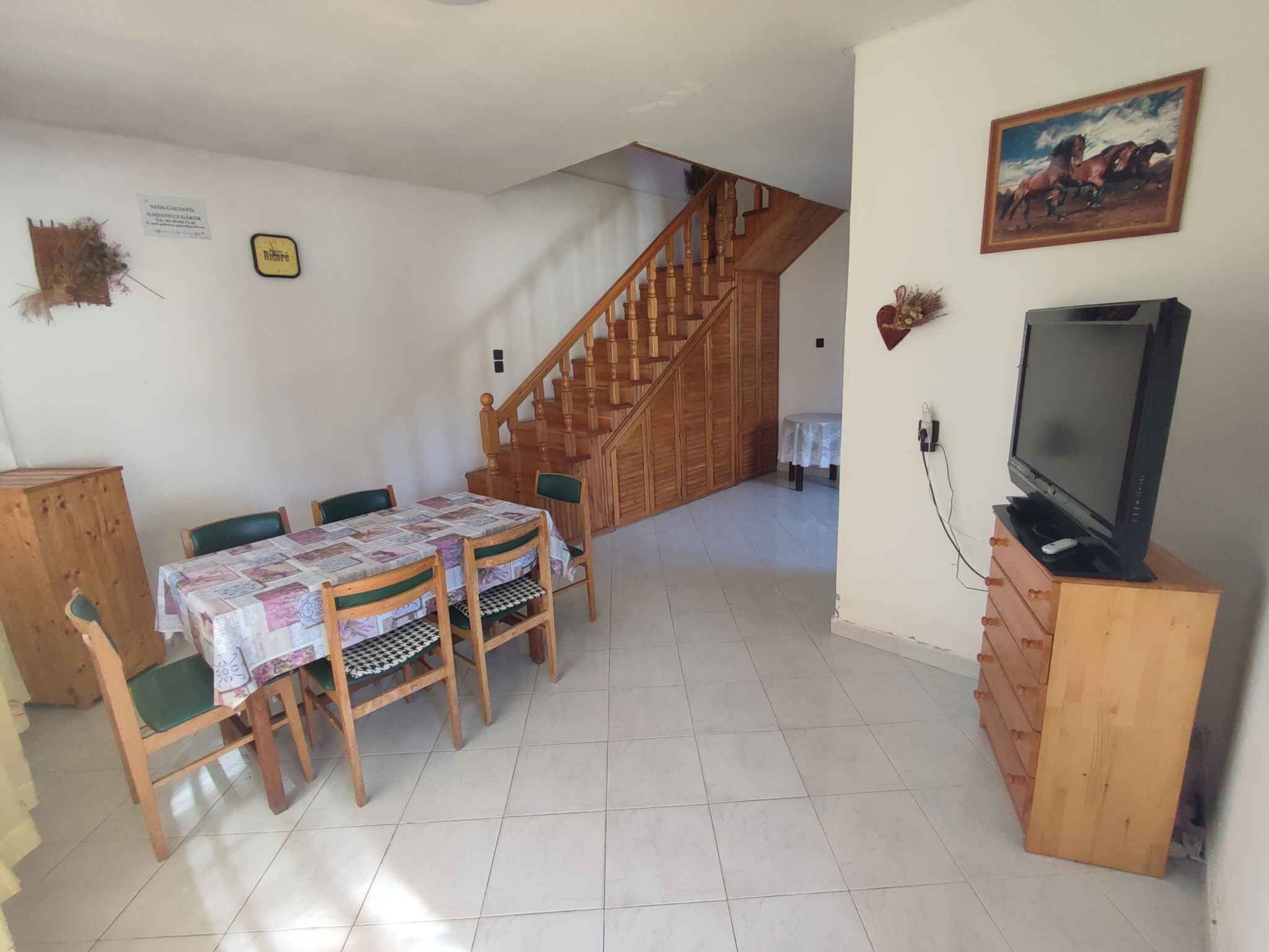 Un coin repas avec une table et des chaises en bois, un escalier, un téléviseur posé sur un meuble en bois et un tableau accroché au mur.