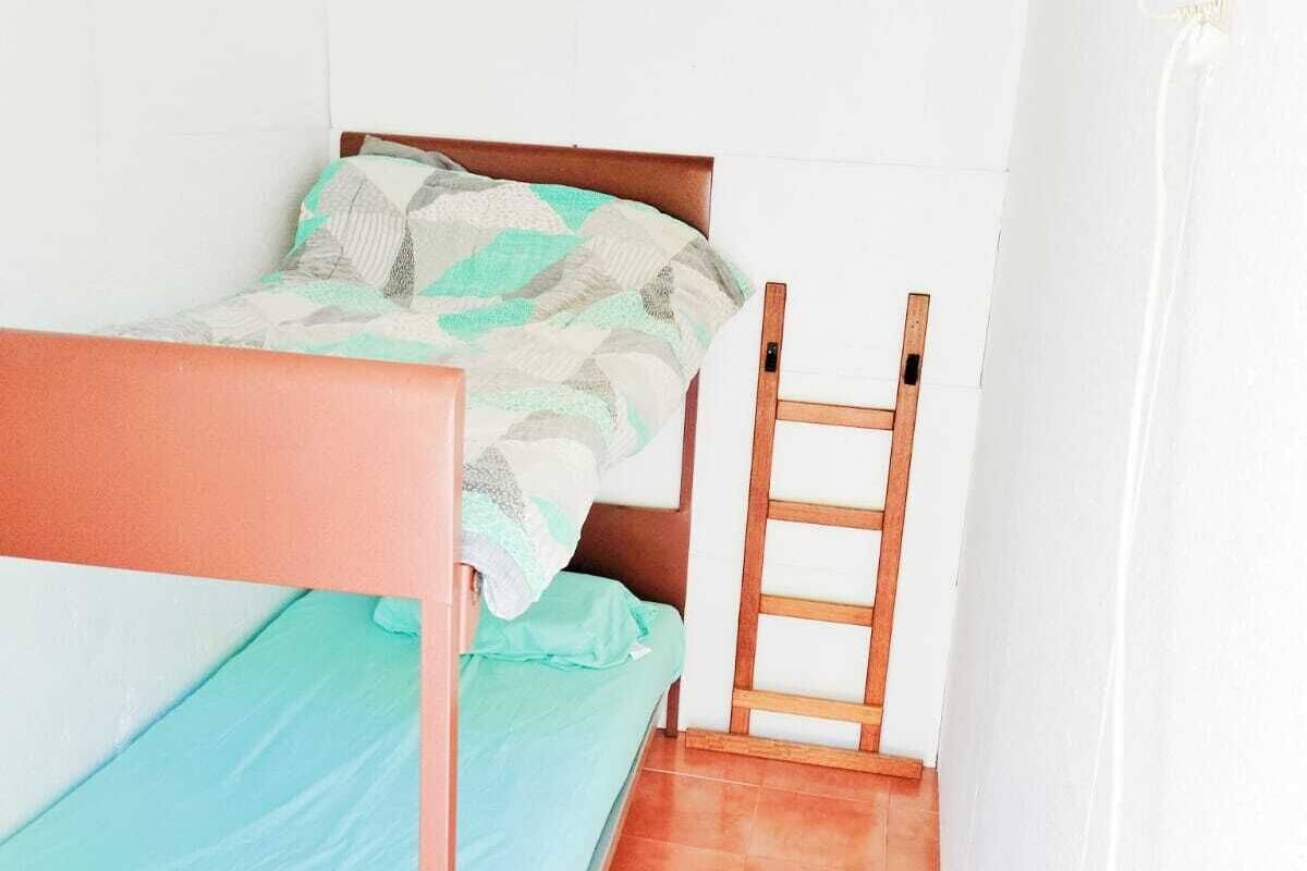 Ein kleines Zimmer mit einem Einzelbett, einem Holzrahmen, einer grünen Bettdecke und einer an der Wand stehenden Leiter.