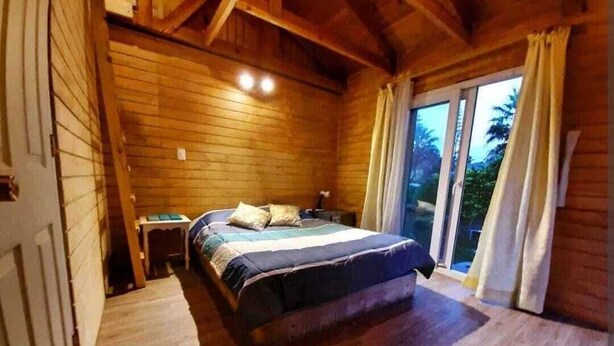 Un dormitorio de cabaña de madera con una cama, una mesita de noche y una ventana con cortinas.