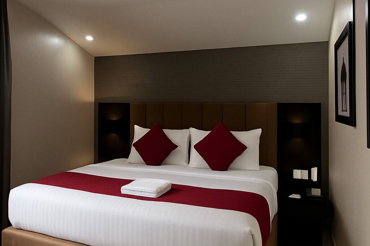 A modern hotel room with a large bed, red and white bedding, a red accent light, a wooden headboard, and a dark nightstand with a lamp.