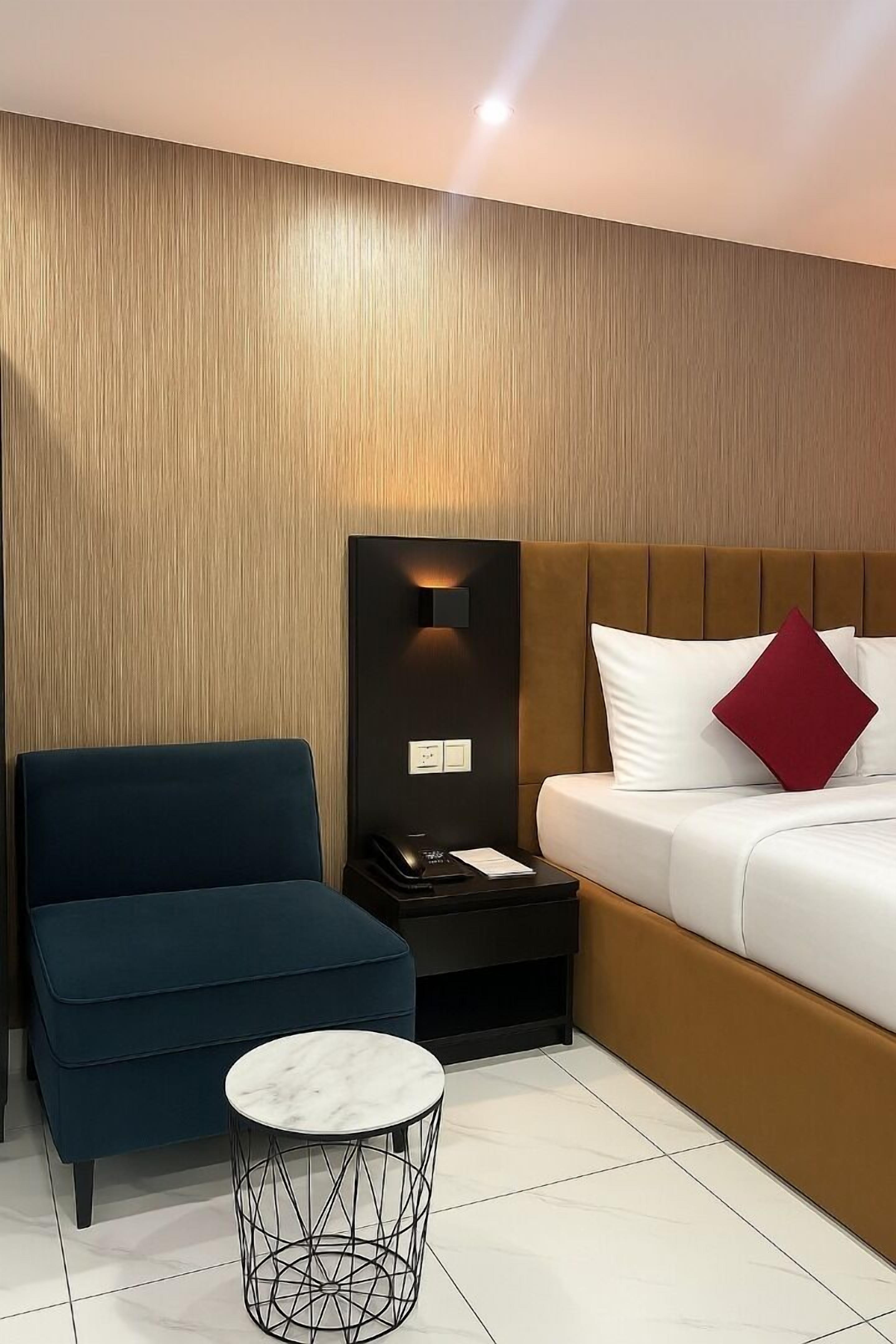 Una habitación de hotel moderna con un panel de pared de madera, un sillón azul, una pequeña mesa redonda y una cama con ropa de cama blanca y una almohada roja.