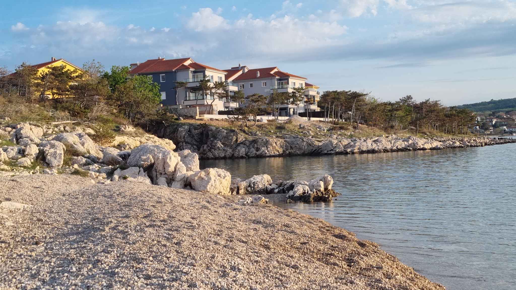 A coastal property with a rocky shoreline, a calm body of water, and a row of modern houses with red roofs.