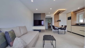 A modern living room with a sofa, a coffee table, and a TV. There is a dining area with a table and chairs. The kitchen is visible with cabinets and a countertop. - Holiday apartment with swimming pool and sea view (Šilo)