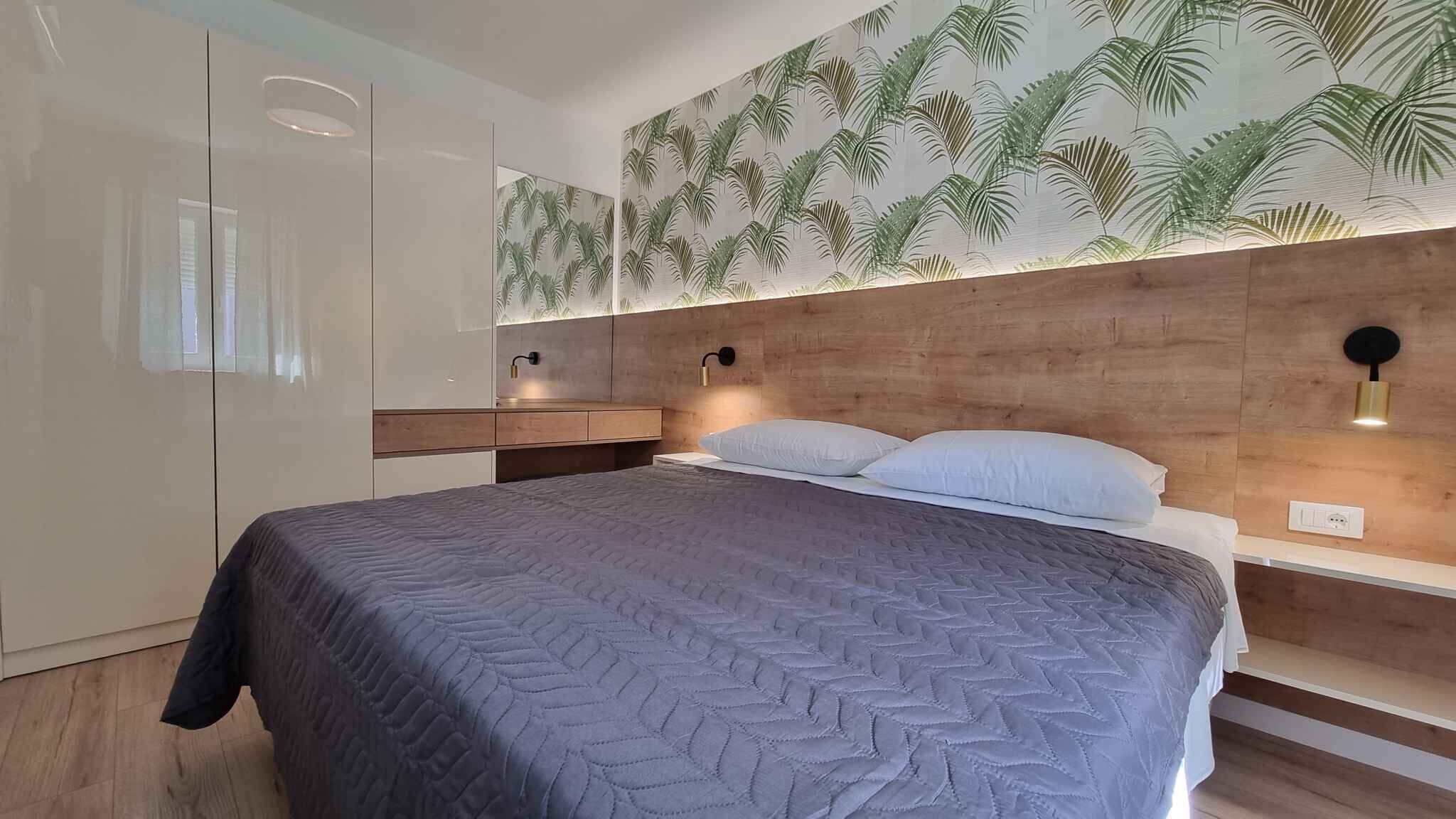 A modern bedroom with a bed, nightstands, a wooden headboard, and a wall with a tropical leaf pattern.
