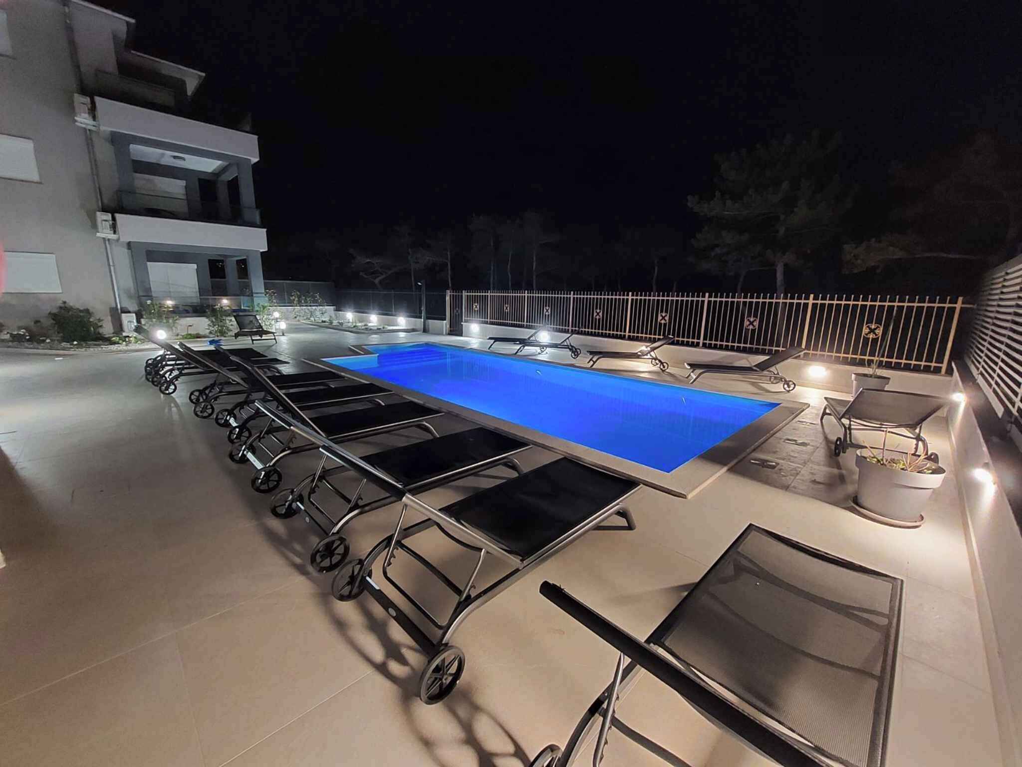 An outdoor swimming pool with lounge chairs and sun loungers, surrounded by a fence and a building.