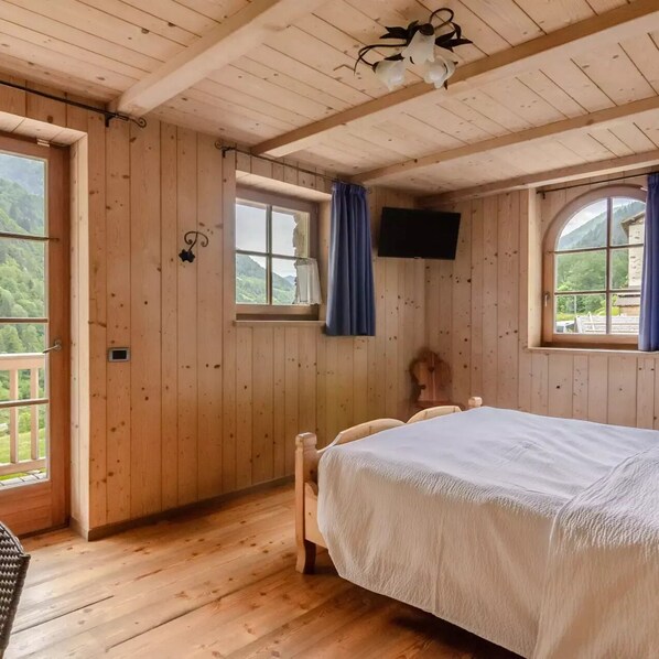 A wooden room with a bed, two windows with curtains, a mounted TV, and a view of greenery outside.
