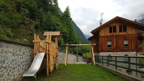 A wooden play structure with a slide and climbing wall, a stone wall, and a wooden building with a sloped roof.