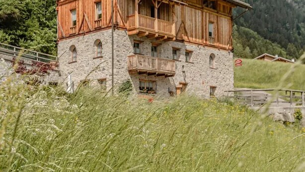 A two-story wooden chalet with stone foundations, set against a mountainous backdrop.