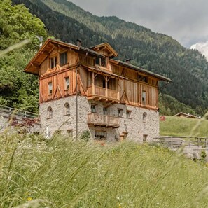 A two-story wooden chalet with stone foundations, set against a mountainous backdrop.