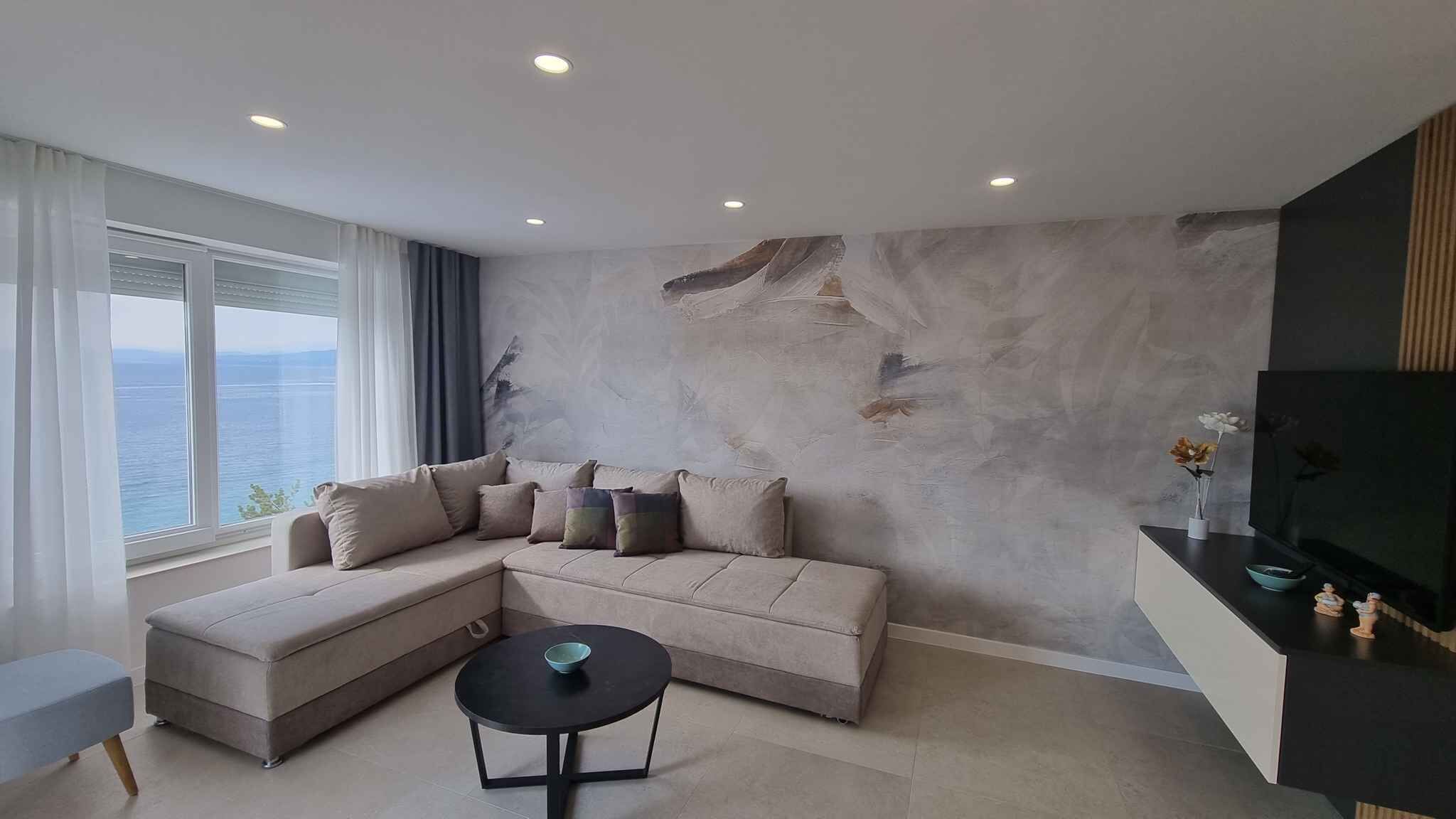Un salon moderne avec un canapé d’angle, une petite table ronde et une vue sur la mer grâce à de grandes fenêtres.