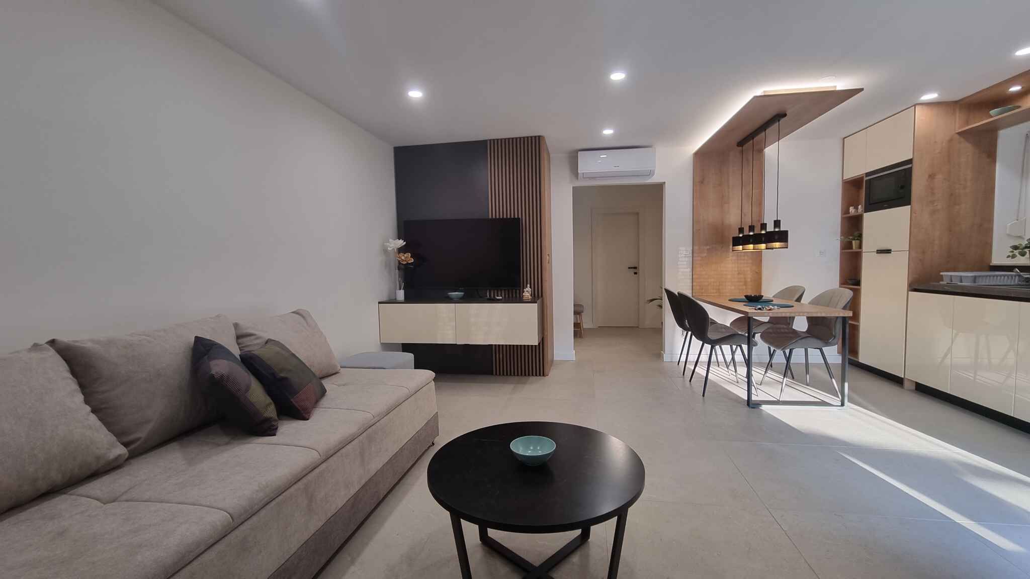 A modern living room with a sofa, a coffee table, and a TV. There's a dining area with a table and chairs. The kitchen has wooden cabinets and a microwave.