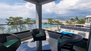 En balkon med fletstole og et glasbord, med udsigt over en pool og et smukt landskab med hav og bjerge.