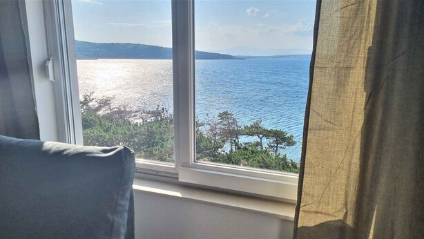 Quarto com uma janela grande que oferece vista para um corpo de água e colinas ao longe.