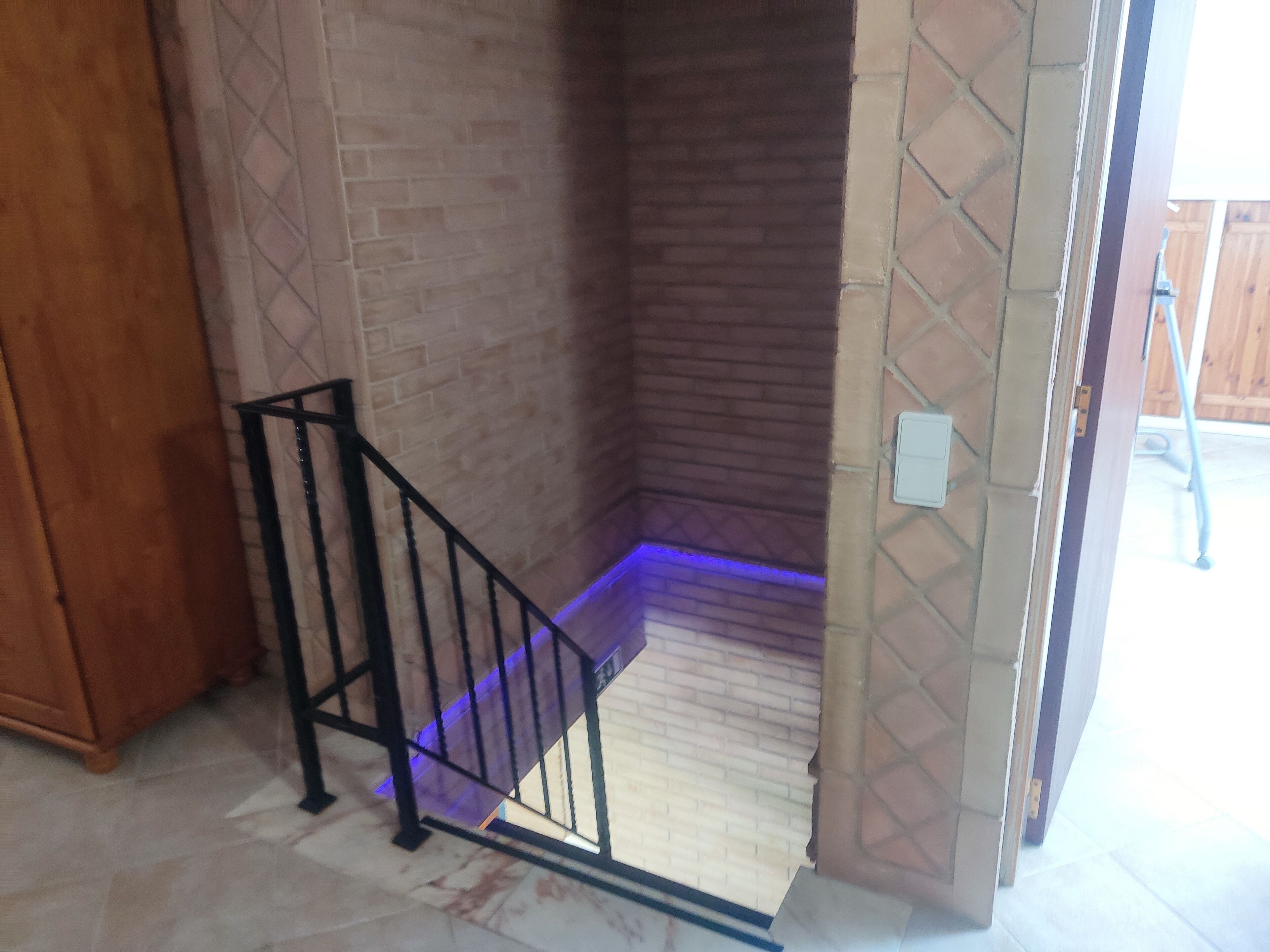 A staircase with a black metal railing leading to a landing. The walls are made of brick and stone, and there is a light switch on the wall to the right.