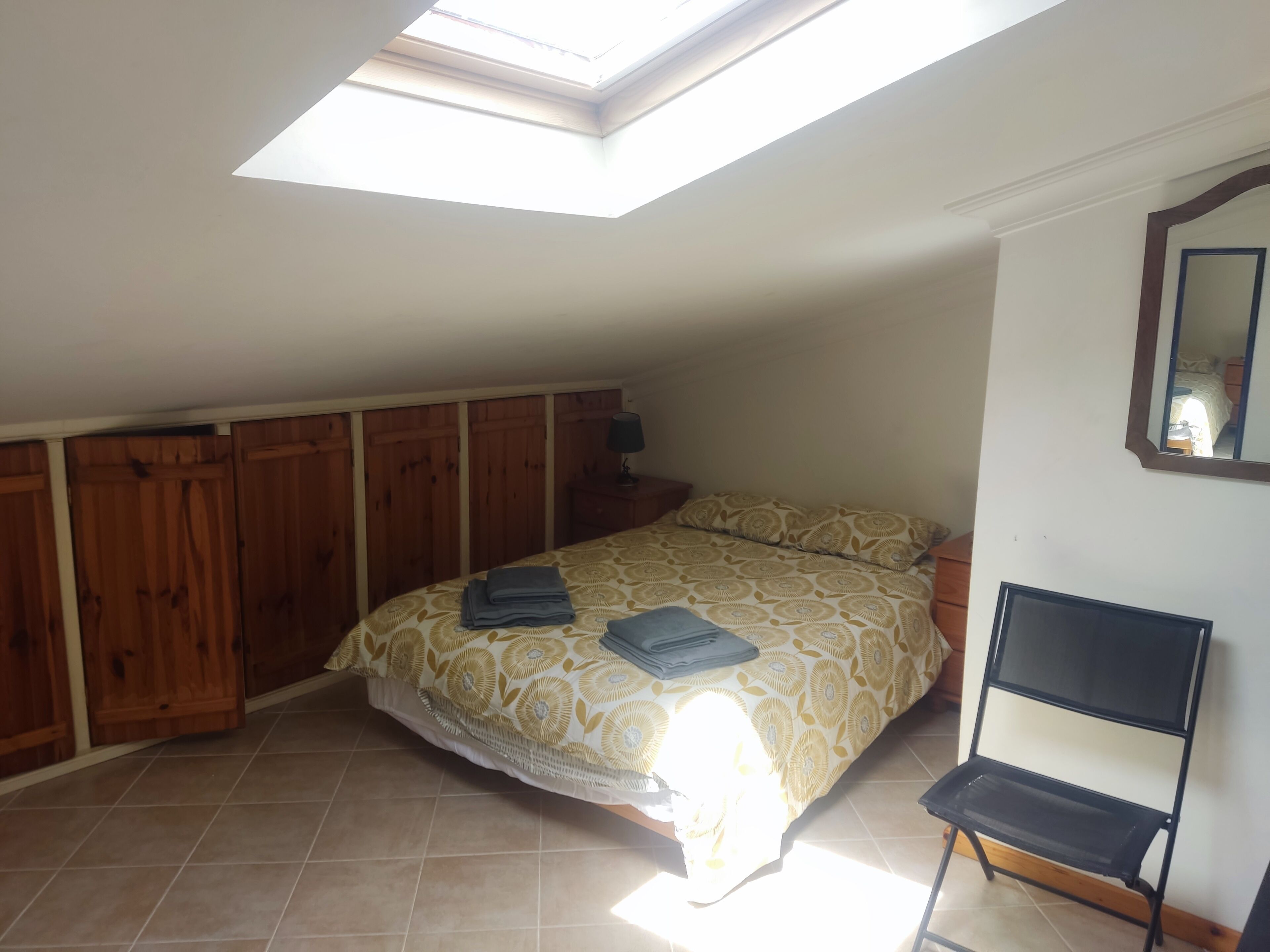 Een slaapkamer met dakraam, een bed met beddengoed met patroon, een stoel en houten lambrisering aan de muren.