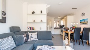 Ein modernes Wohnzimmer mit einer grauen Eckcouch, einem Couchtisch mit einer Zeitschrift und einer Kerze sowie einem Essbereich mit einem Holztisch und Stühlen.