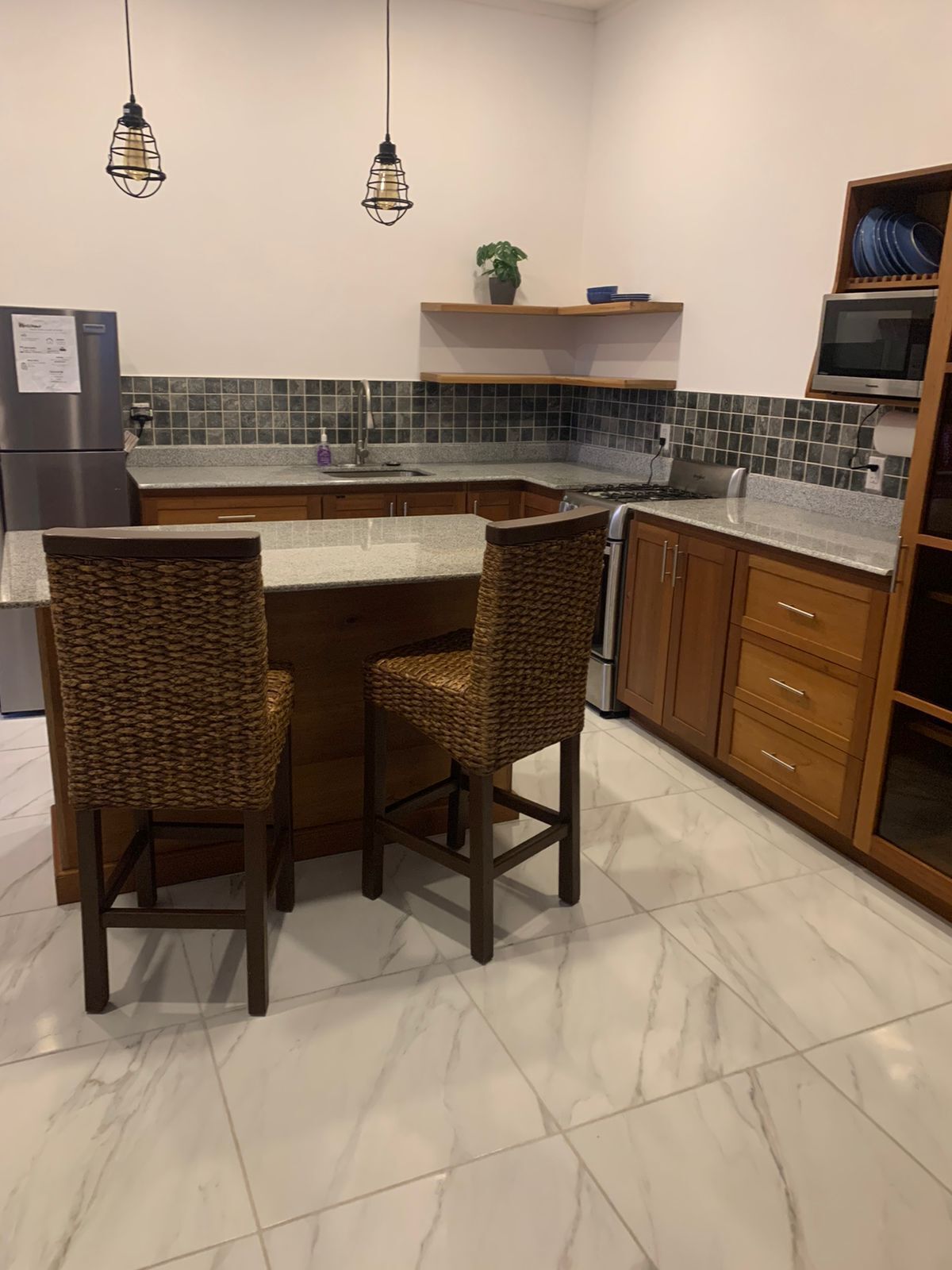 A kitchen with a marble countertop, two wicker bar stools, a stainless steel refrigerator, a microwave, and a sink.