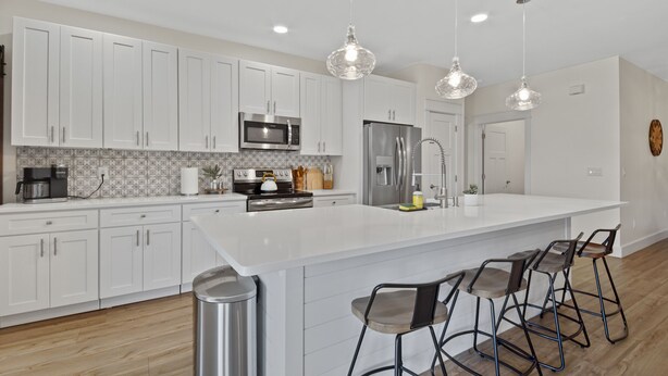 Una cocina moderna con armarios blancos, electrodomésticos de acero inoxidable, una isla grande con taburetes altos e iluminación colgante.