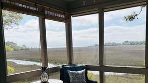 Une véranda grillagée, meublée de meubles en osier, dotée d’un ventilateur de plafond et offrant une vue sur un paysage naturel.