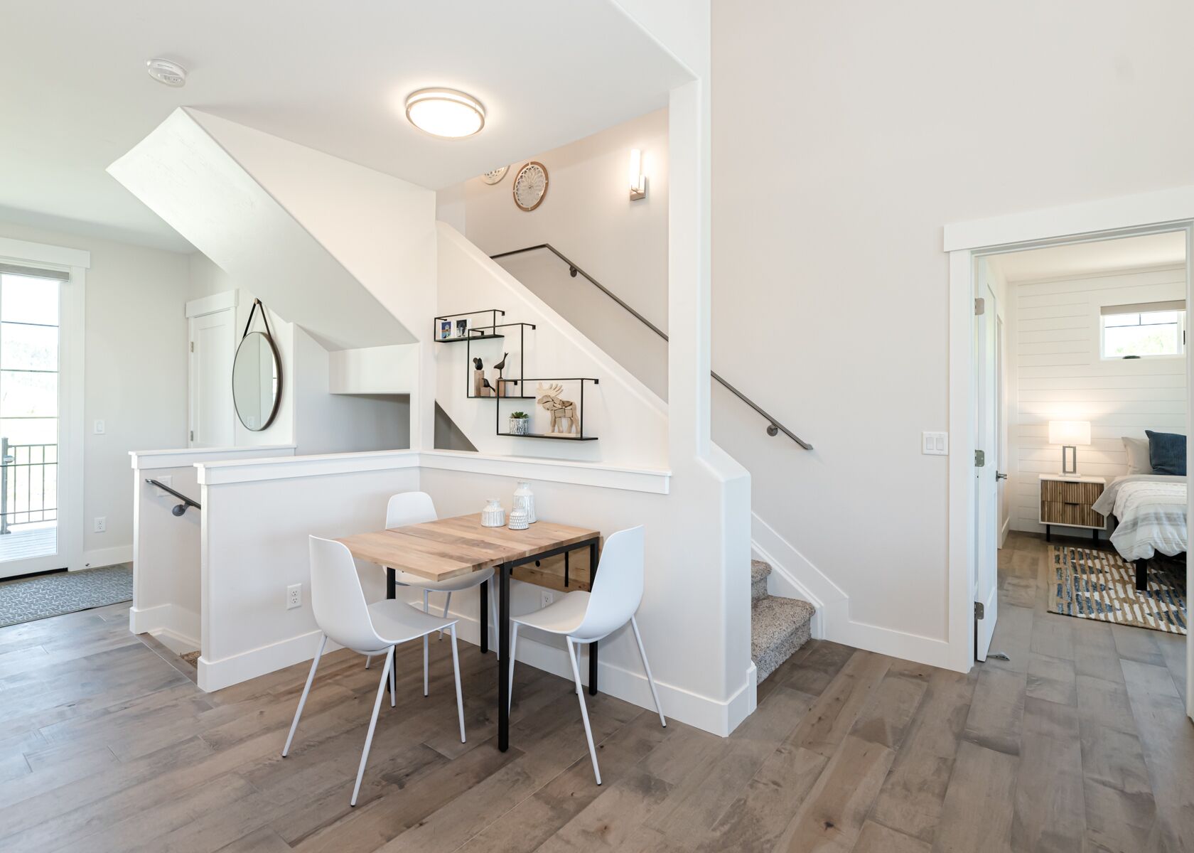 A modern kitchen with a wooden table and white chairs, a staircase with a railing, and a dining area with a view to the outside.