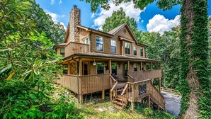 A two-story wooden house with a stone chimney, wooden deck, and a staircase leading to the porch.