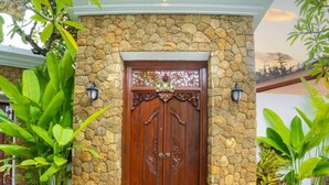 A stone facade with a wooden double door, flanked by two wall-mounted lights, and surrounded by lush greenery. - Alam Jepun Villa  (Legian)
