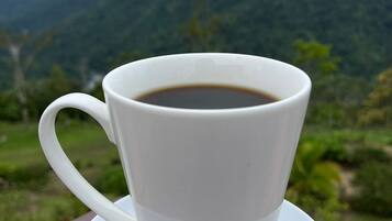 A white cup of coffee on a white saucer, placed on a balcony with a view of green hills and a cloudy sky.