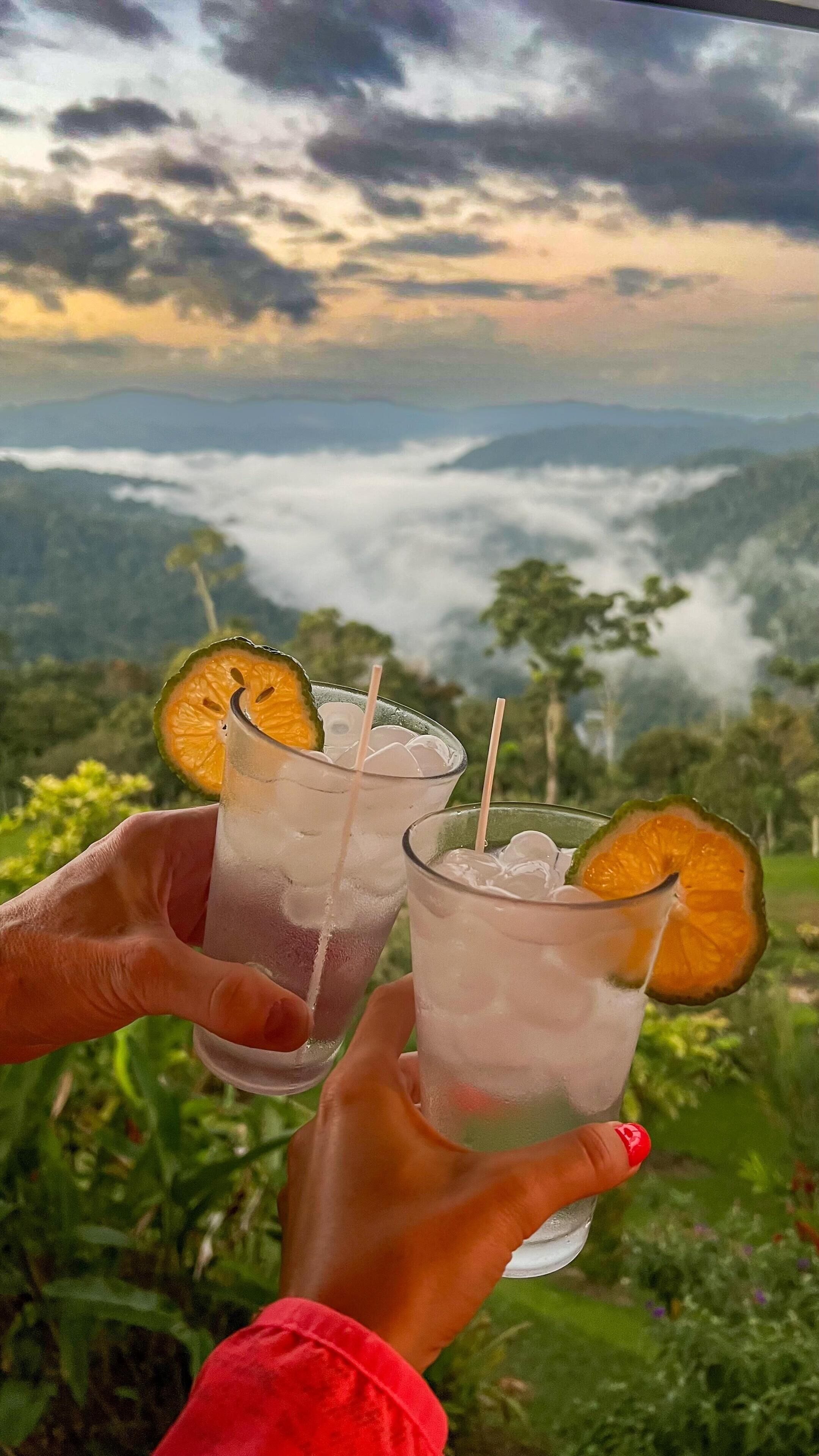 Dos vasos de una bebida con hielo y una rodaja de naranja, sostenidos por manos sobre un paisaje montañoso y un cielo nublado.
