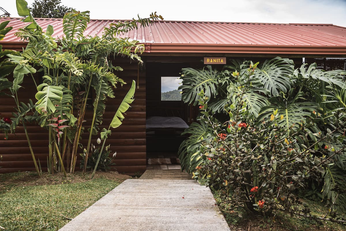A wooden cabin with a red roof, a sign reading "RANITA", and a concrete pathway leading to the entrance.