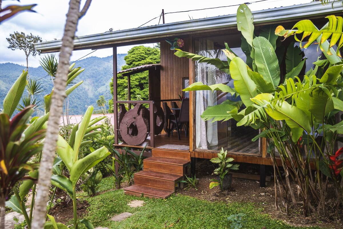 A wooden house with a porch, surrounded by lush greenery and a mountain backdrop.